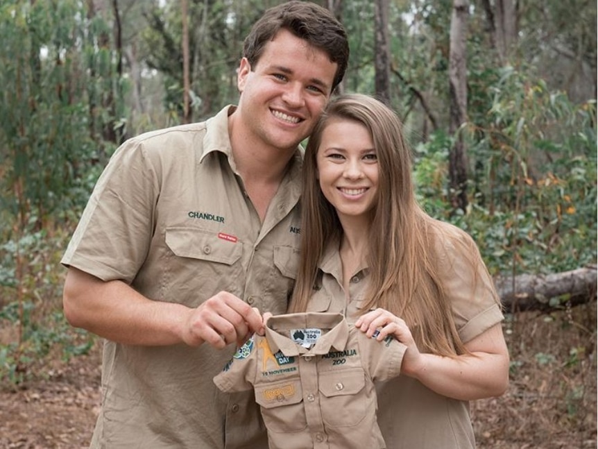 Two people in khaki fatigues hold up a baby shirt