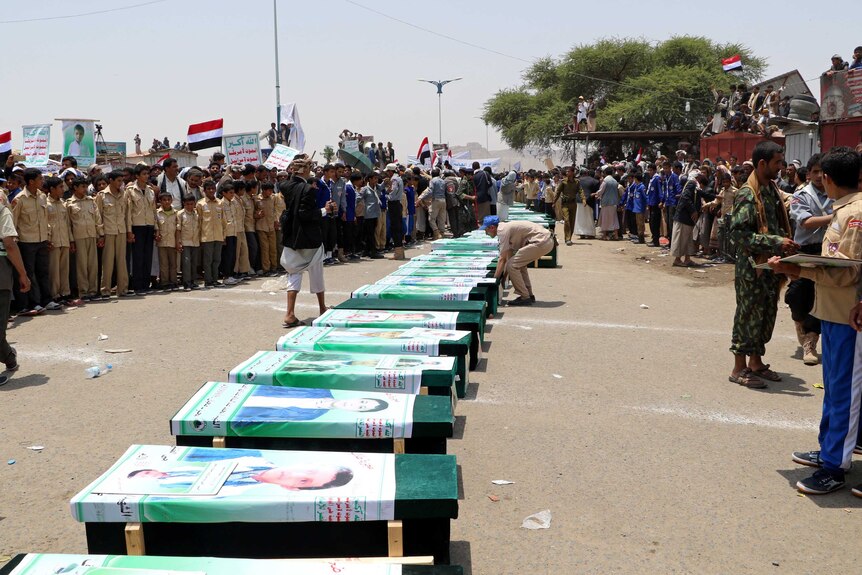 A crowd gathers in front of a row of coffins with pictures of children stuck on them.