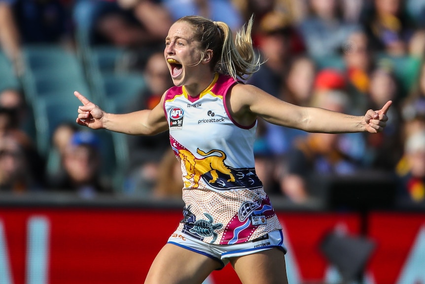 A Brisbane Lions AFLW player runs with her arms out wide as she celebrates a goal.