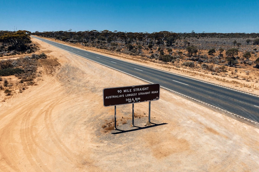 A sign alongside a straight flat country highway, pale peach coloured earth, blue skies.
