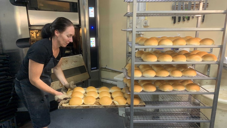 A woman wearing an apron puts trays of bread buns onto a large cooling rack.