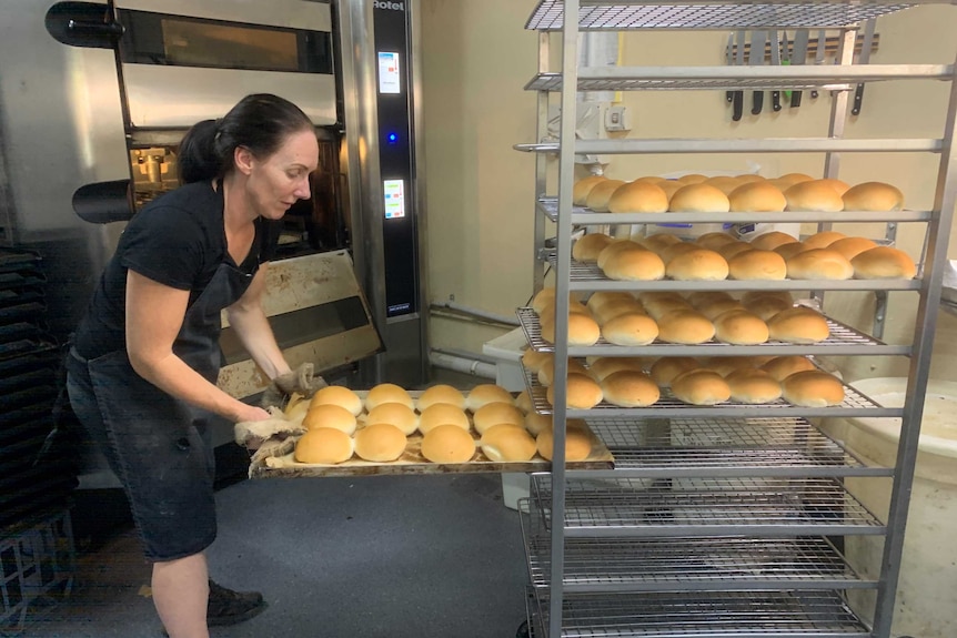 A woman wearing an apron puts trays of bread buns onto a large cooling rack.