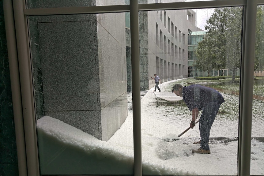 Large piles of hail are dug into by people at Parliament house, seen through the window.