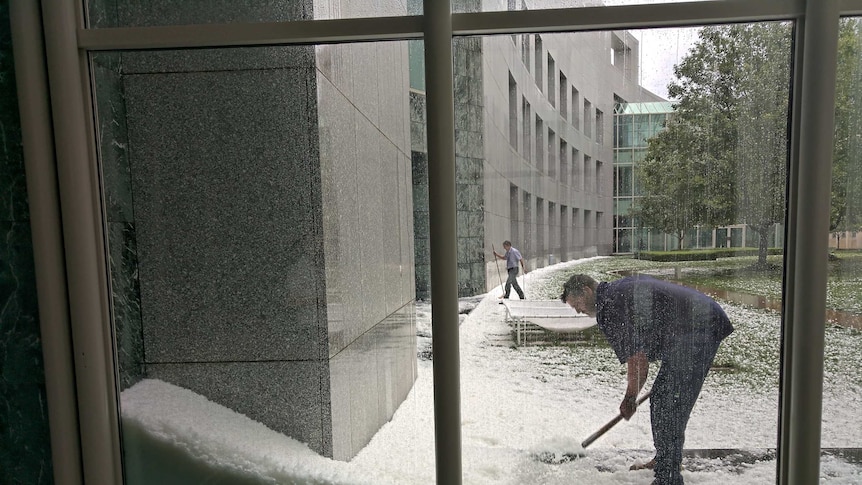 Large piles of hail are dug into by people at Parliament house, seen through the window.