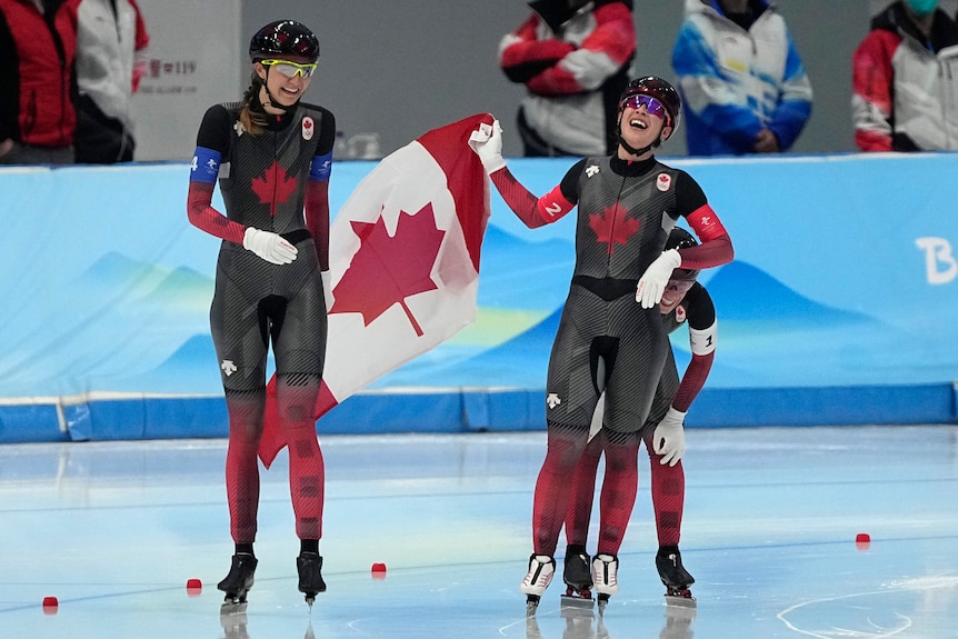 Canadian speed skaters smile and breathe in as they wave the Canadian flag in celebration of a gold medal.