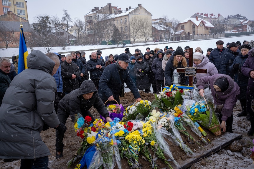 Peopl place bunches of wrapped flowers on a fresh grave while it snows.