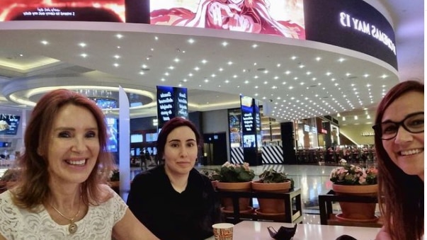 Three women sit at a table at a brightly lit mall with a restaurant in the background.