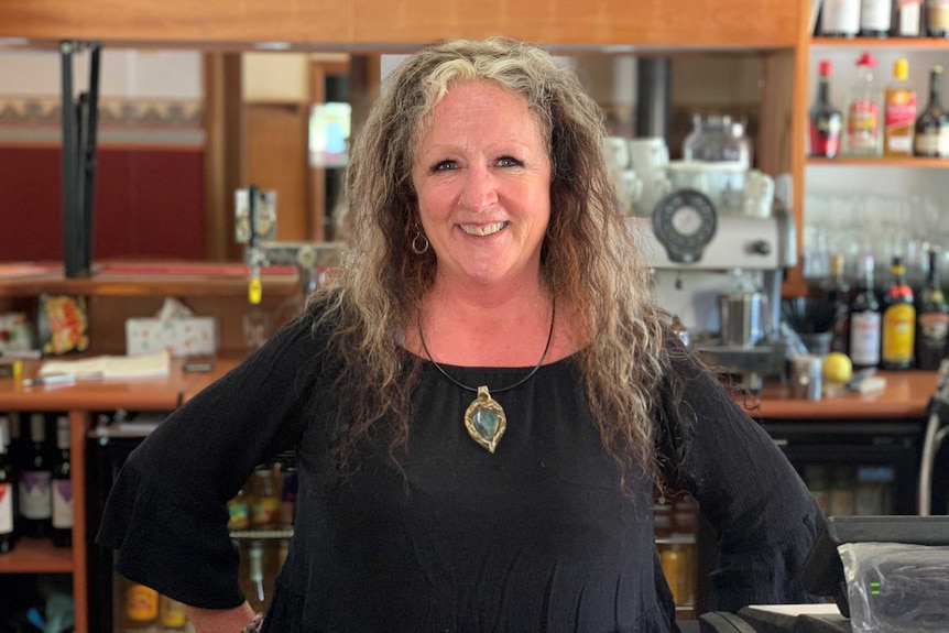 A middle-aged woman with a kind smile. She has blonde and brown, wild hair, and faint eyebrows. She's standing in a pub.