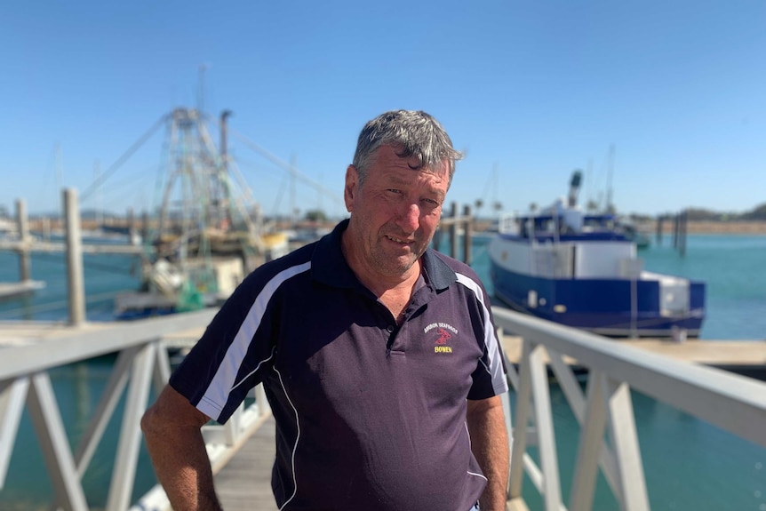 A man stands in front of a marina with fishing boats in the background