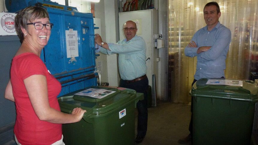 Two people with large green bins next to a man holding a blue machine.