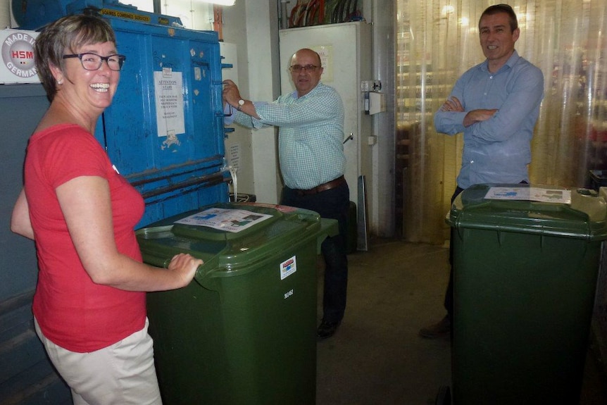 Two people with large green bins next to a man holding a blue machine.