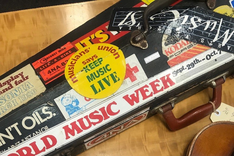 A closed violin case is covered with stickers. The violin sits on a table next to the case with a letter in between.