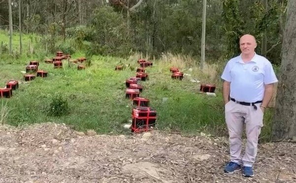 Man standing next to a collection of red bee hives on grass