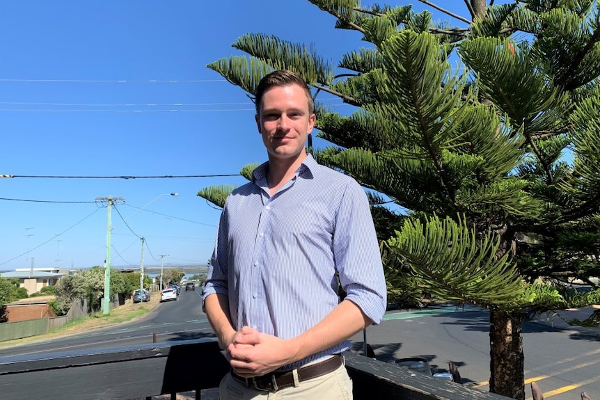 A young man wearing a pale blue business shirt stands on a street in front of a large pine tree on a sunny day.