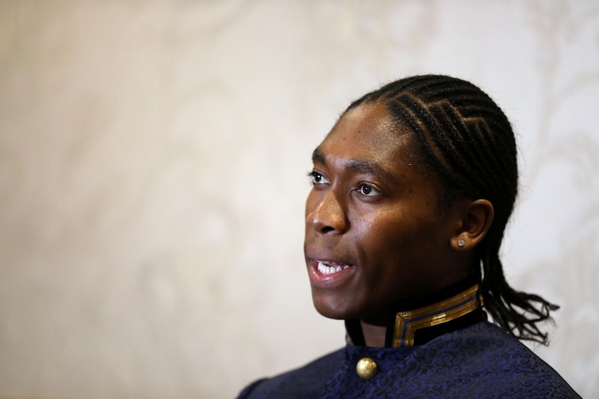 A young black woman with braided hair speaks in front of an off-white wall.