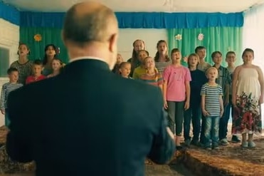 A man in a suit has his back to the camera and looks towards a group of children standing in lines wearing coloured clothes