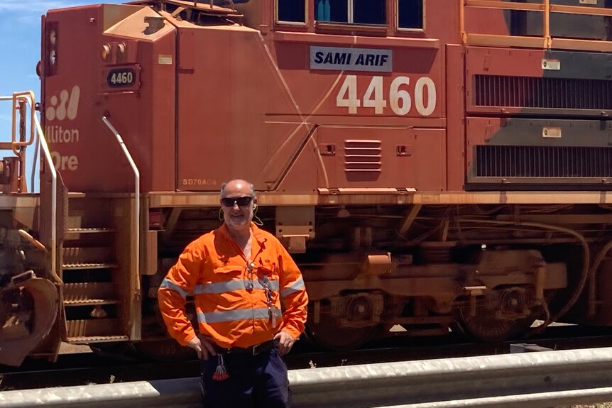 A man in high visibility clothing stands with his hands on his hips in front of an iron ore train.