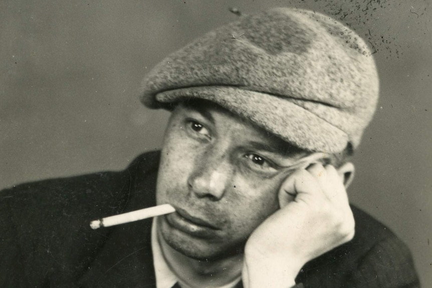 A young man rests his head on his hand with a cigarette in his mouth.