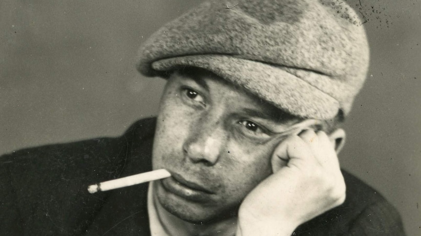 A young man rests his head on his hand with a cigarette in his mouth.
