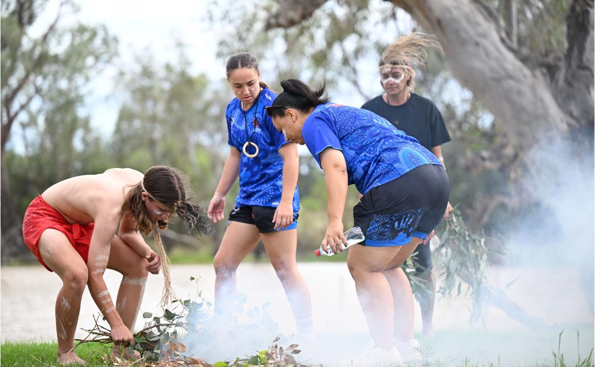 An Aboriginal man in traditional dress puts leaves on a small fire while three women watch.
