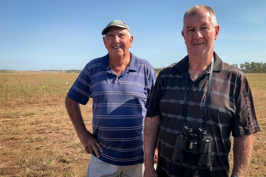 Two older men, one wearing a cap and one with binoculars, stand in a large field of dry grass next to Darwin airport.