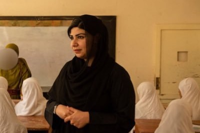 A woman stands in a classroom, she is wearing a black hijab.