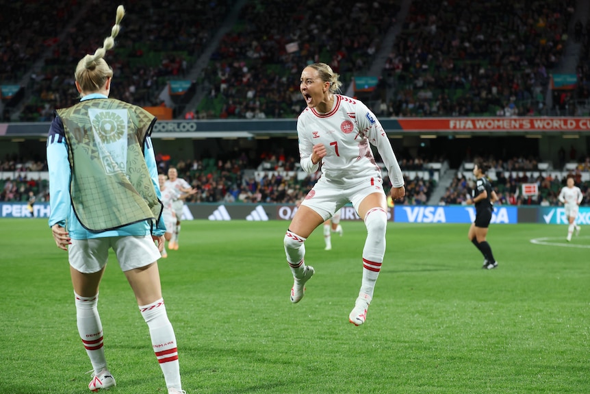 A Danish footballer jumps in the air in celebration, as her teammate runs with hair flying after a goal.