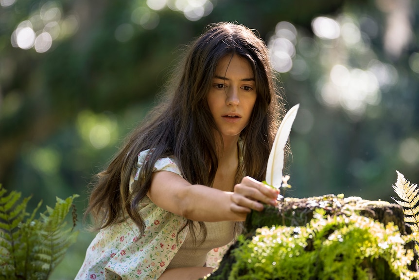 A long-haired brunette reaches out to grasp a white feather, with an entranced expression