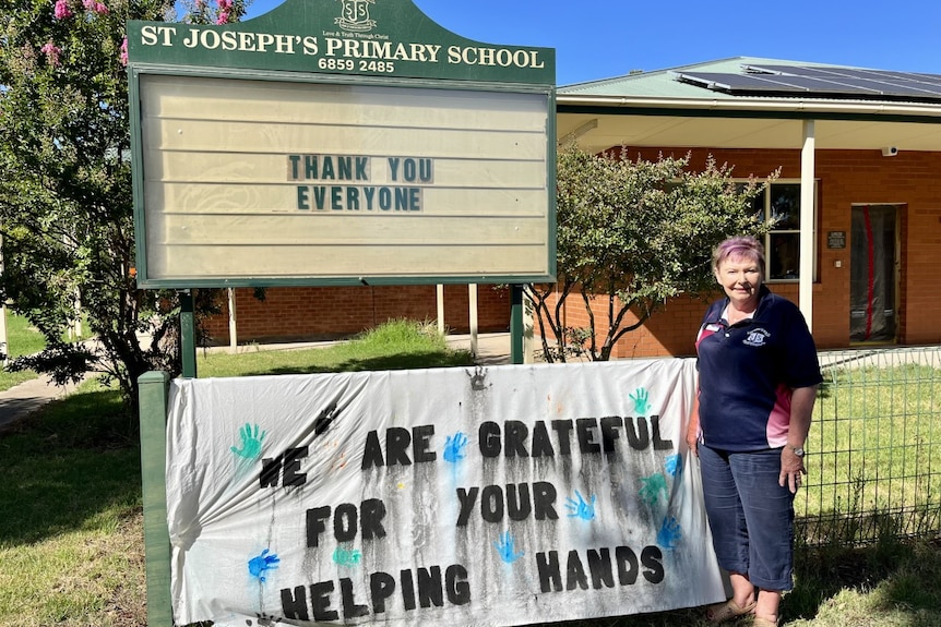 A woman standing in front of the entrance to a school next to a sign saying: thank you everyone, we are grateful for your help