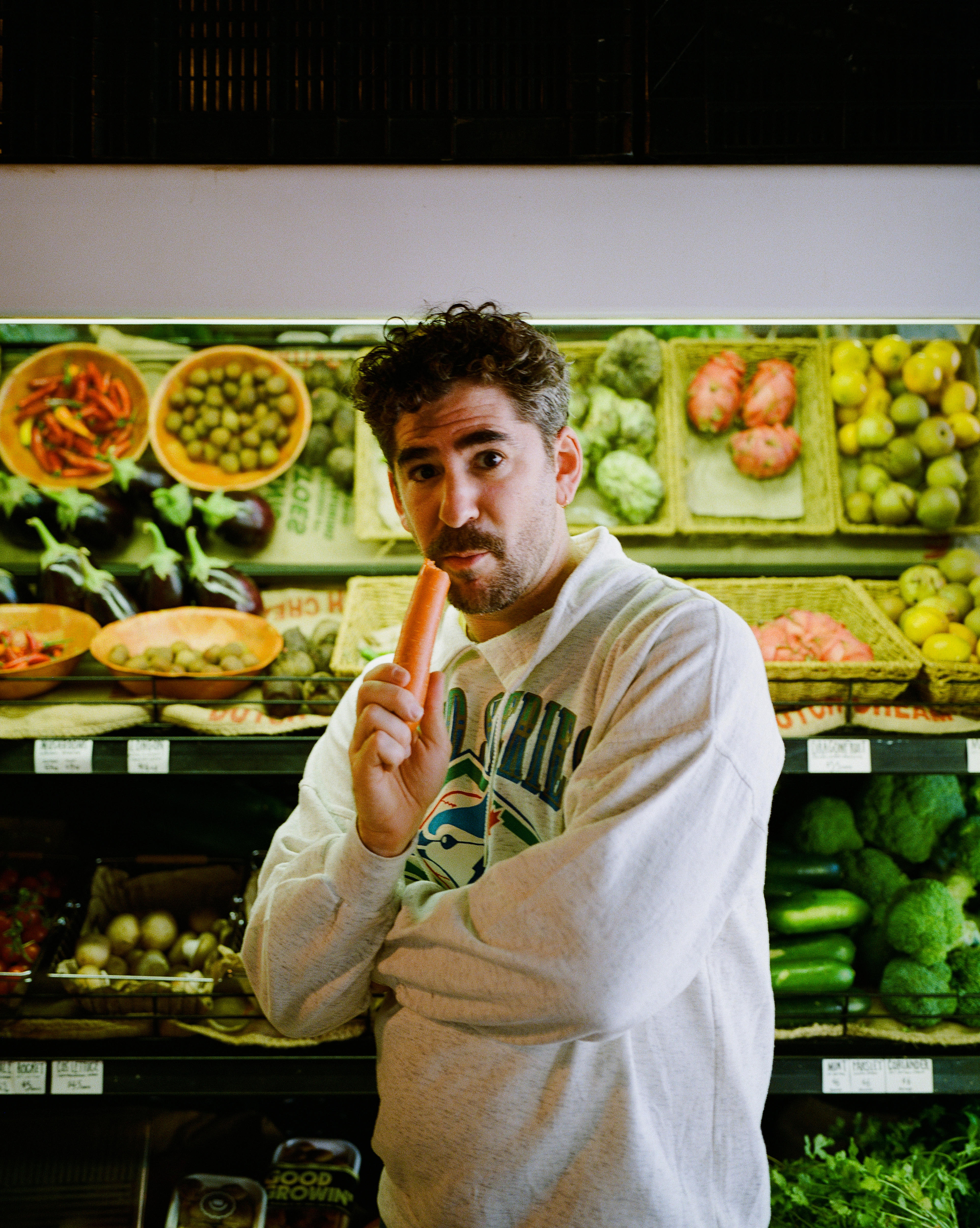 Um homem está na seção de produtos hortifrutigranjeiros de um supermercado, segurando uma cenoura nos lábios.