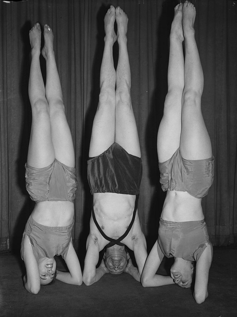 Black and white retro image of Sir Paul Dukes doing a handstand on his head alongside two ballerinas.