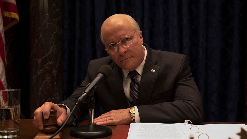 Colour still of Christian Bale looking solemn while seated in front of microphone and his hand on a gavel in 2018 film Vice.