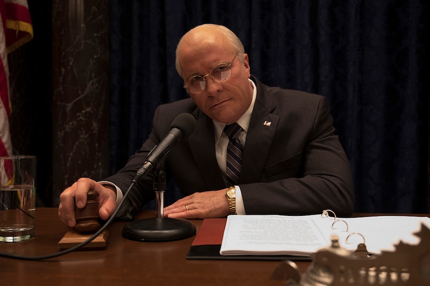 Colour still of Christian Bale looking solemn while seated in front of microphone and his hand on a gavel in 2018 film Vice.
