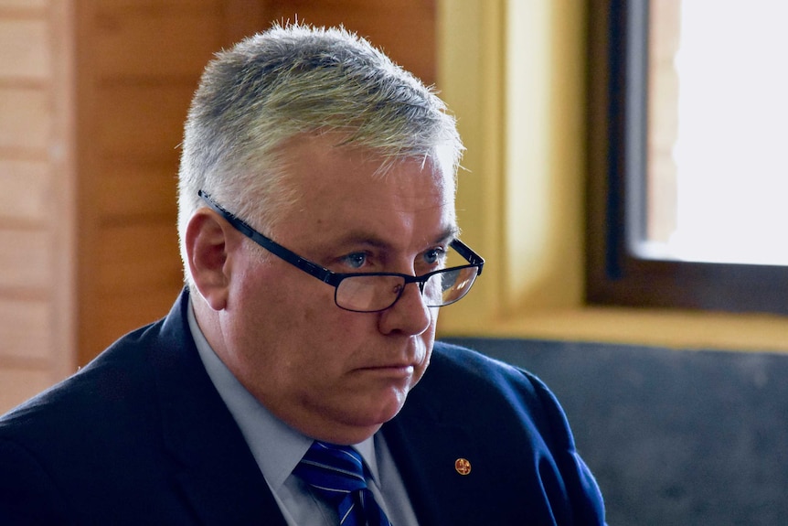 A man with white hair and glasses stares off camera.  He is wearing a blue suit.