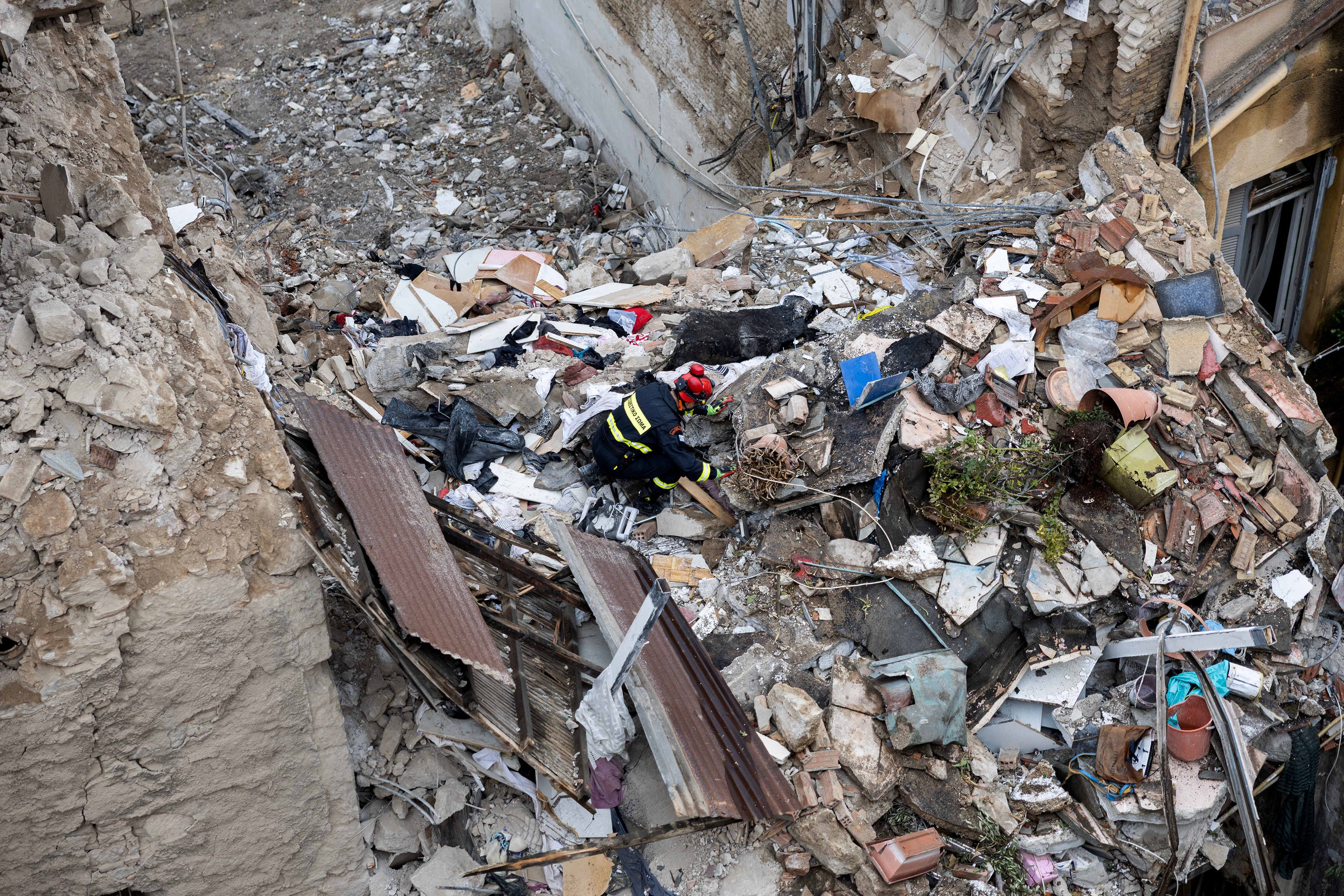 A firefighter in a red helmet and black overalls seen on top of a large pile of rubble, cluttered by concrete rocks.