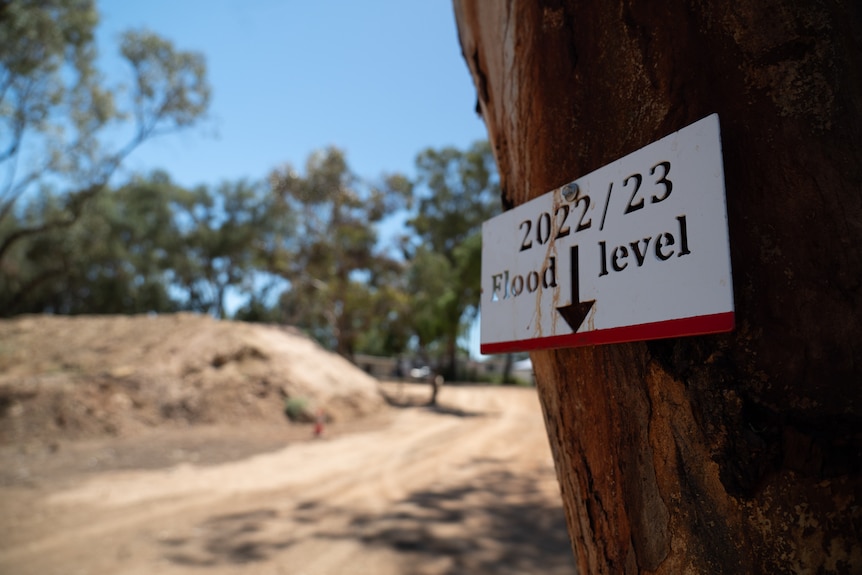 A red and white sign reads '2022/23 flood level' with an arrow pointing down to a red line, a dirt levee is behind it