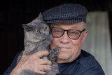 An older man in a cap stands on his front door step holding a small blue-grey cat