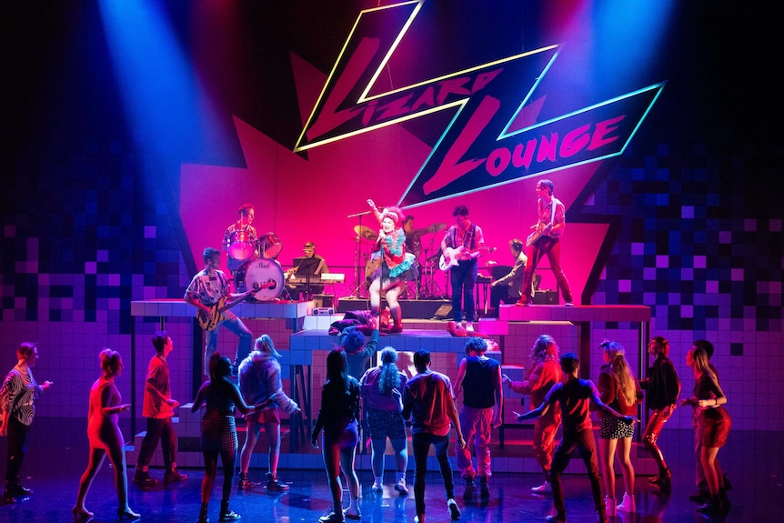 A stage filled with a group of young people dressed in 1980s costumes, a band performing for a crowd, in the musical Starstruck