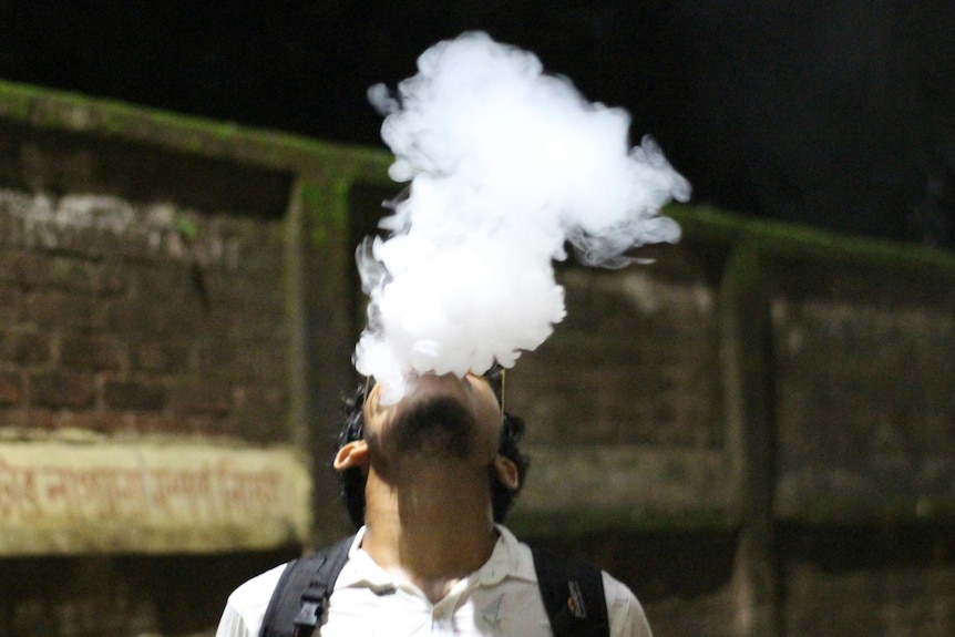 A man vaping tilts his head upwards as a plume of smoke rises from his mouth and nose.