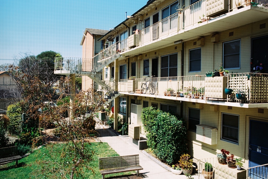 On a bright day, you view a 1960s-era triple story block of flats with balcony access on the outside of the structures.