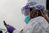 A healthcare worker with long hair and full PPE looks at a tablet at a testing site.