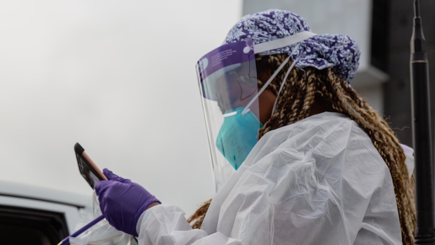 A healthcare worker with long hair and full PPE looks at a tablet at a testing site.