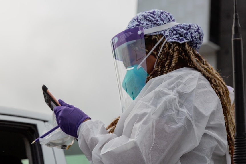 A healthcare worker with long hair and full PPE looks at a tablet at a testing site.