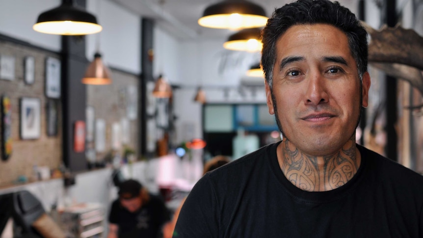A smiling Maori man wearing a black shirt stands in a Sydney tattoo shop.
