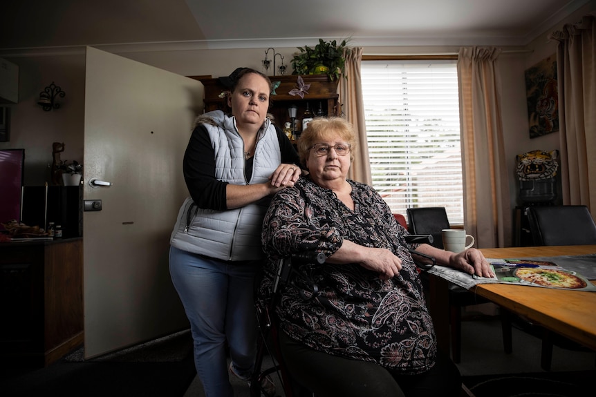 A woman sits at a dining table with a younger woman standing next to her chair.