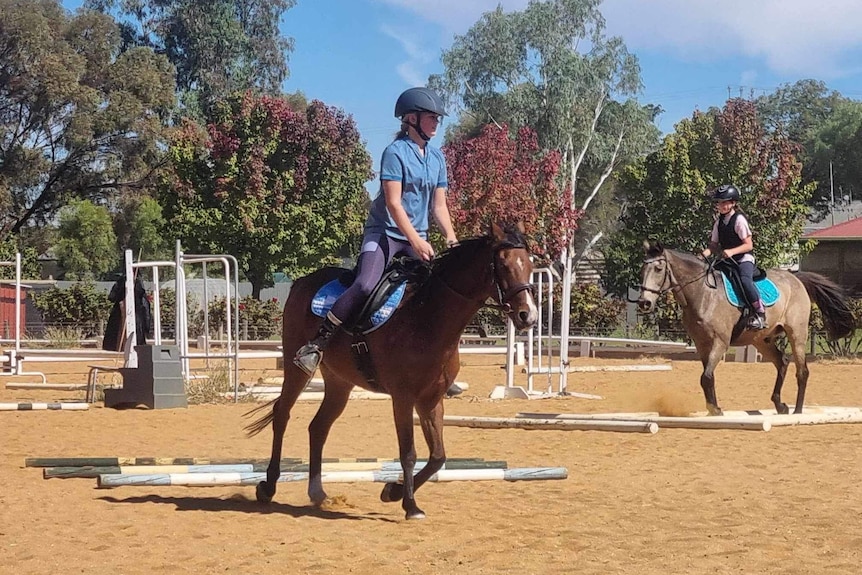 A girl in a blue shirt rides a brown horse around equestrian jumping obstacles, another girl rides a light brown horse behind.