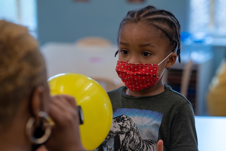 A child in a red mask reaches for a yellow balloon