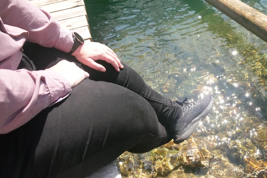 Feet in black sneekers and black jeans hanging over jetty above shallow water at the ocean edge.