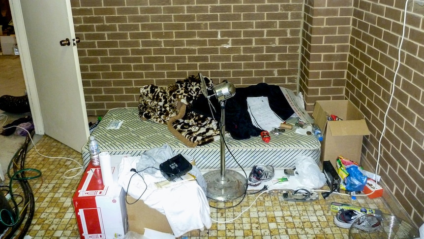 A floor mattress in a brown brick room with a microwave on the floor and general personal items.