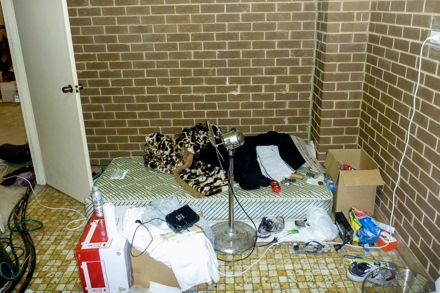 A floor mattress in a brown brick room with a microwave on the floor and general personal items.